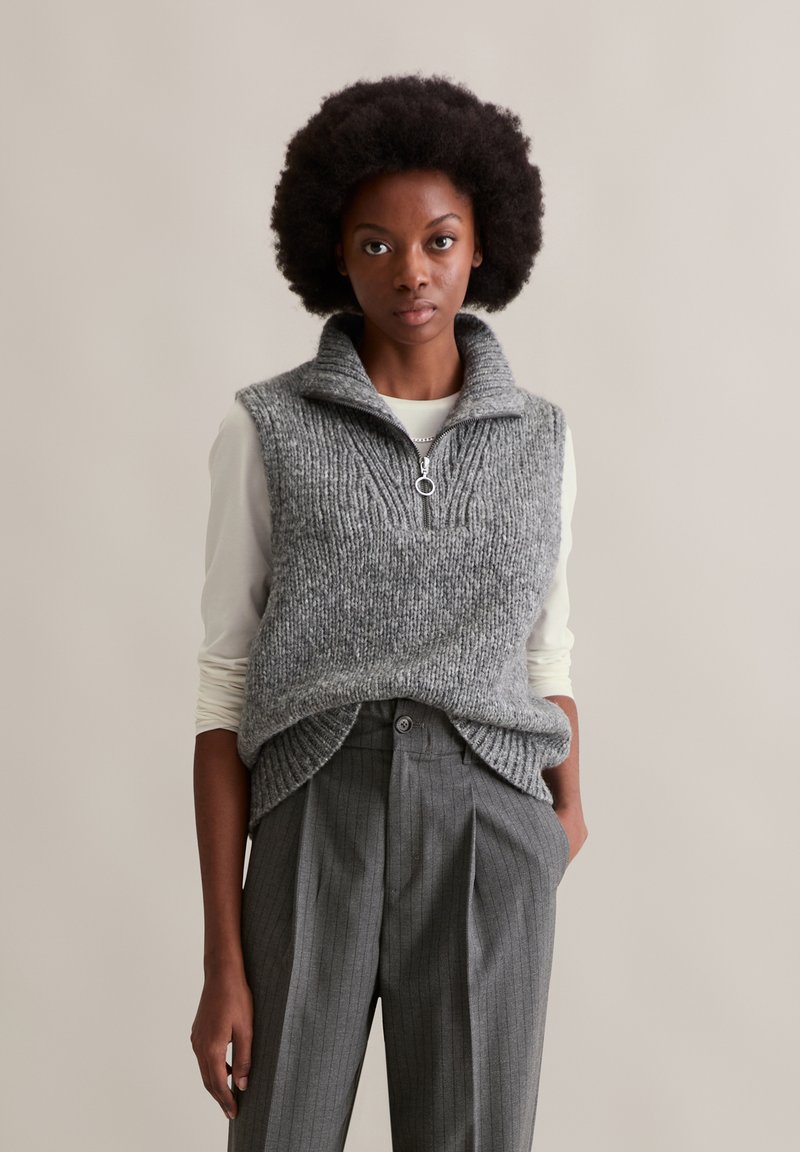 Frau mit Afro-Frisur, die einen grauen, gestrickten ärmellosen Pullover mit Reißverschluss und graue Nadelstreifenhosen vor einem beigen Hintergrund trägt.