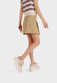 Beżowa spódnica plisowana zestawiona z krótką, wzorzystą bluzką i jasnymi sneakersami z metalicznymi akcentami oraz okrągłym noskiem.