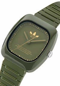 Kvadratisk olivgrön handledsklocka med matchande rem, guldfärgade visare och markeringar, Adidas treklöverlogotyp och slogantext på urtavlan.