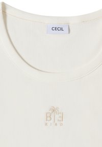 Top blanc côtelé à col rond avec une petite broderie beige d'un palmier et les lettres « BEE KINO » sous l'étiquette de la marque « CECIL » à l'intérieur du col.