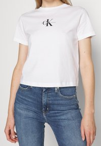 Bílý bavlněný zkrácený tričko s černým logem Calvin Klein Jeans na hrudi. V kombinaci s tmavě modrými přiléhavými džíny.