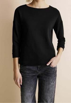 Osoba ubrana w czarną, teksturowaną bluzkę z długim rękawem i wyblakłe czarne dżinsy, z jedną ręką w kieszeni, na jednolitym beżowym tle.