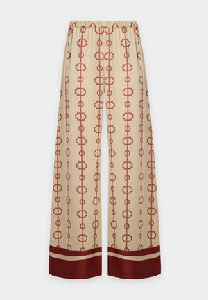 Frau mit langen, glatten Haaren, die ein lockeres schwarzes Off-Shoulder-Top und weite beige Hosen mit rotem geometrischem Muster trägt.