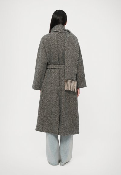 Manteau long en chevrons gris avec un col et une ceinture, avec un détail de foulard à franges. Associé à un jean bleu clair ample.