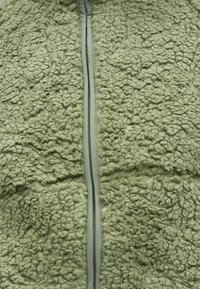 Fleecová bunda v jemně zelené barvě, s texturovaným povrchem, zapínáním na zip a hladkým lemováním podél zipu. Kapsy nejsou viditelné.