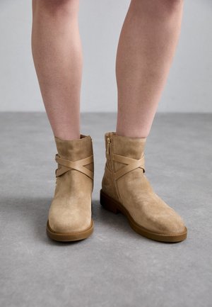 Paar hellbraune Wildleder-Stiefeletten mit Kreuzriemen und seitlichen Reißverschlüssen, getragen an nackten Beinen vor grauem Boden.