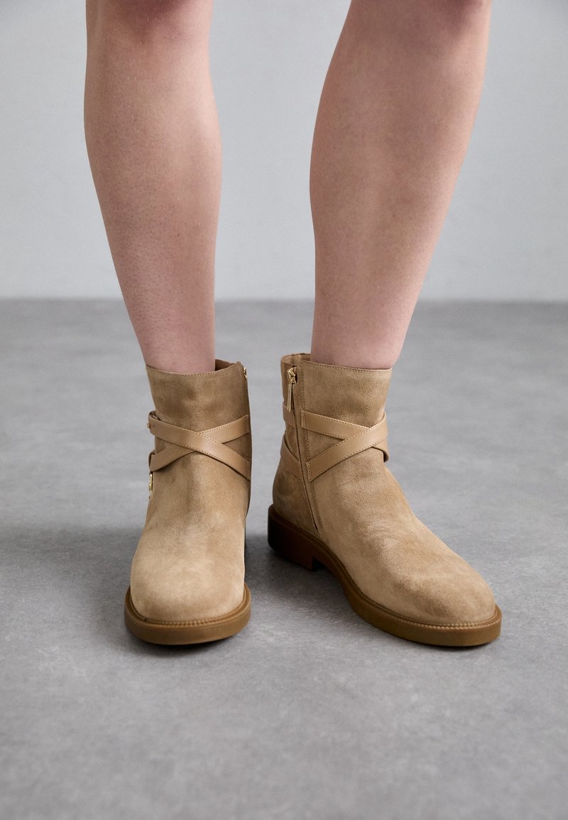 Paire de bottines en daim beige avec brides croisées et fermetures éclair latérales portées sur jambes nues sur fond de sol gris.