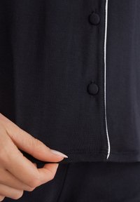 Zwart katoenen top met een slim fit, voorzien van een witte bies en ronde knopen. Close-up toont een hand die de onderrand vasthoudt, waardoor de textuur van de stof wordt benadrukt.