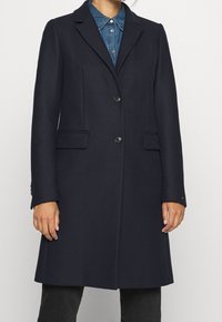 Marineblauwe wollen blend jas met omgeslagen revers, twee voorzakken, twee knopen en een getailleerde pasvorm, gecombineerd met een denim overhemd eronder.