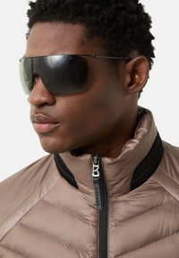 Zonnebril met grote, donkere lenzen, zwart metalen montuur, gedragen met een lichtbruine donsjas met rits bij de hals en mesh accenten.