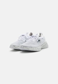 Chaussures de sport blanches avec une tige en maille texturée, des lacets plats et une semelle en caoutchouc tachetée. Présentent un logo sur le côté et un design minimaliste.