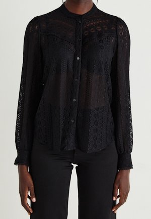 Femme portant un chemisier noir en dentelle à manches longues avec un motif floral et un pantalon noir, posant devant un fond uni.