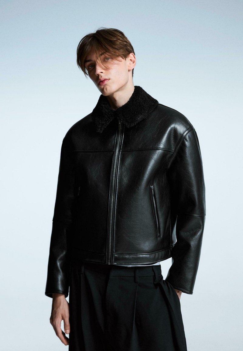 Crna kožna jakna s mekim crnim ovratnikom od janjeće kože, zatvaranjem na rajsferšlus i bočnim džepovima. Dizajn je strukturiran i moderan.