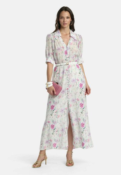 Femme portant une robe maxi blanche à fleurs avec des manches bouffantes, une taille ceinturée, tenant une pochette rose, accessoirisée avec des bracelets blancs et des talons dorés.