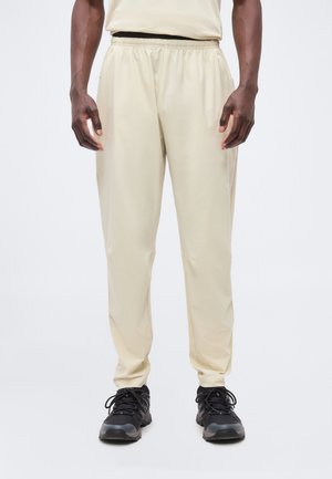 Hombre que lleva pantalones beige con cintura elástica y zapatillas deportivas negras, de pie frente a un fondo liso y claro.