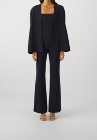 Femme portant un blazer-cape noir sur une combinaison noire à jambes évasées et des sandales à talons couleur chair, debout devant un fond uni clair.