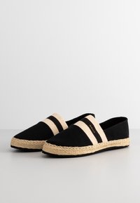 Espadrilles noires en toile avec deux rayures beiges, semelle en jute, et un design plat. Détails du logo en texte sur le côté.