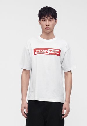 Joven con una camiseta blanca de Diesel con logo rojo, de pie frente a un fondo claro y simple, mirando al frente con una expresión neutra.