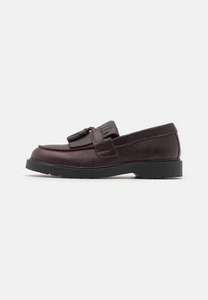 Mocassin en cuir marron foncé avec une surface texturée, des glands sur le dessus, et une semelle en caoutchouc noire sur un fond blanc.