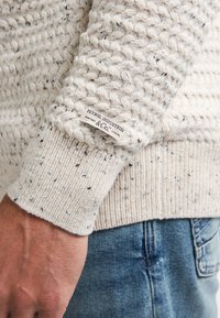 Beige gebreide trui met een gestructureerd steekpatroon en geribbelde mouwen. Etiket op de mouw zegt "Petrol Industries".