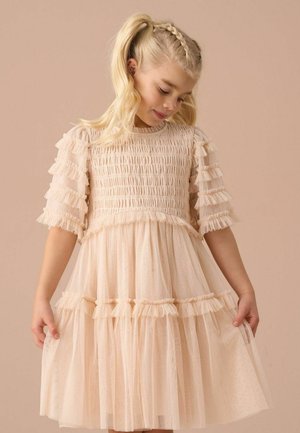 Junges Mädchen mit blonden Zöpfen, trägt ein hellrosa Kleid mit Rüschen an Ärmeln und Rock, schaut nach unten und hält die Seiten des Kleides.