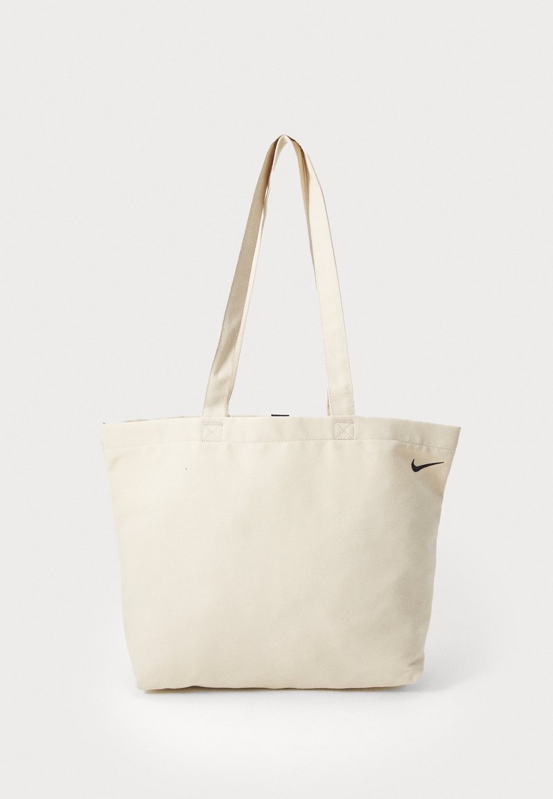 Borsa a spalla in tela beige con due lunghe maniglie, dotata di un piccolo logo Nike nero su un lato. Design semplice e rettangolare con cuciture a vista.