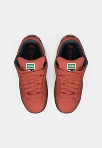 Červené semišové tenisky s okrúhlou špičkou, viazaním a polstrovaným interiérom. Obsahujú zelené logo Puma a čierny akcent na vložke.