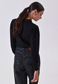 Top nero a maniche lunghe con spalle a palloncino indossato con jeans neri a vita alta con dettagli in paillettes, creando un look stratificato e texturizzato.