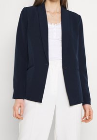 Kvinde iført navyblå blazer, hvid tanktop med blonder, hvide højttaljede bukser og guldfarvet vedhængsvedhæng, stående foran en ensfarvet baggrund.