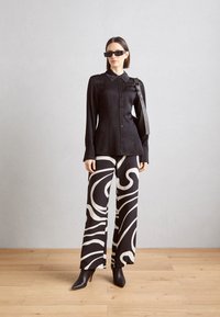Zwarte langarm button-up shirt met een kraag, gecombineerd met zwart-wit gestreepte wijde broek. Model draagt zwarte zonnebril en hoge hakken.