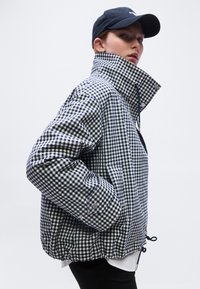 Άτομο με καρό μπλε ναυτικό και λευκό σακάκι με ψηλό γιακά και μαύρο καπέλο, ποζάρει με τα χέρια στις τσέπες μπροστά σε απλό φόντο.