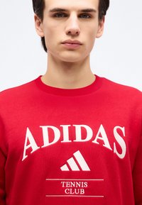 Jeune homme portant un sweat-shirt rouge Adidas avec le texte blanc "Tennis Club" et le logo sur le devant, sur un fond clair uni.