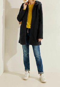 Abrigo negro con cremallera y capucha, de textura suave, combinado con una camiseta amarilla de manga larga, vaqueros azules y zapatillas blancas. Detalles incluyen bolsillos.