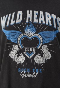 T-shirt noir avec un motif graphique bleu et blanc comprenant un cœur, des ailes, des roses, et le texte "WILD HEARTS CLUB ROCK THE WORLD."