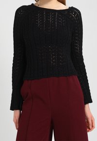 Donna che indossa un top in crochet nero a maniche lunghe e pantaloni bordeaux a vita alta e gamba larga su uno sfondo semplice.