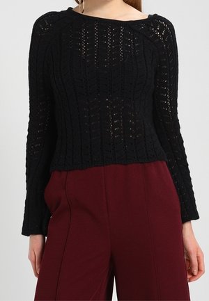 Woman wearing a black long-sleeve crochet top and high-waisted wide-leg burgundy pants against a plain background.