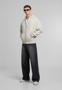 Lys beige zip-up hettegenser med frontlomme, parret med vide grå denimbukser og hvite sneakers. Modell med solbriller.
