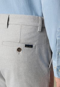 Lichtgrijze wollen broek met een achterzak met knoop en een blauw label, gedragen met een lichtblauw overhemd met lange mouwen.