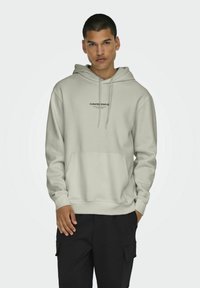 Lys grå hettegenser med snøring, en frontlomme i kenguru-stil, og svart tekst som sier "KURERT HEBLAR." Stoffet ser jevnt og mykt ut.