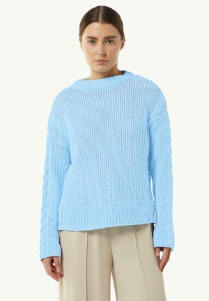 Light blue knitted sweater with a ribbed texture, wide neckline, and textured cable details on the sleeves, paired with cream-colored trousers.