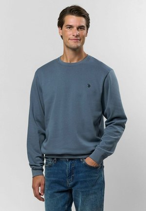 Modrá mikina vyrobená z mäkkého materiálu, s okrúhlym výstrihom, dlhými rukávmi a malým čiernym logom na hrudi. Spárovaná s džínsami.