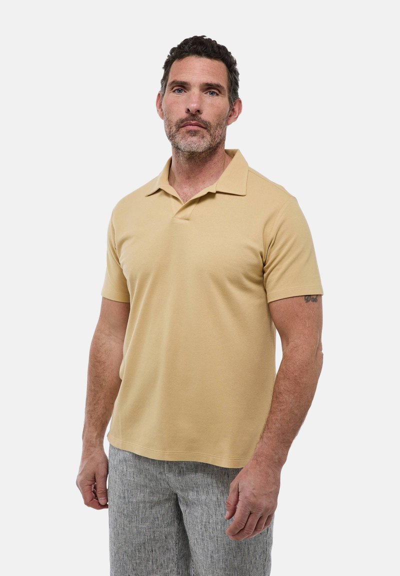 Polo à manches courtes de couleur beige clair, avec un col classique et un tissu lisse et texturé. Porté avec un pantalon à rayures claires.