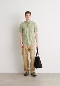 Camisa de botones de manga corta en verde claro, pantalones beige, zapatillas verdes y un bolso tote negro. Diseño simple con un corte relajado.
