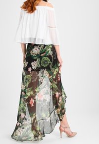 Fließender schwarzer Maxi-Rock mit floralen Motiven in Form von grünen Blättern und rosa Blumen, mit gestuften Rüschen und transparenter Stofftextur. Kombiniert mit nude-farbenen High Heels.