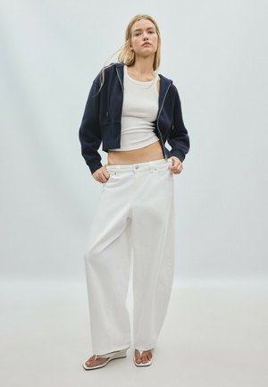 Femme blonde portant un débardeur blanc, un sweat à capuche court bleu marine, un pantalon blanc oversize et des tongs blanches, debout devant un fond uni.