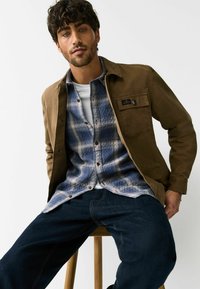 Barna dzseki kék kockás flanel ing fölött, fehér környakas póló alatt, sötét farmernadrággal párosítva. A modell egy fa széken ül.