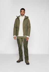 Giacca isolante verde oliva con cappuccio, abbinata a un maglione lavorato a maglia crema e pantaloni cargo coordinati. Stivali da escursionismo neri e arancioni.
