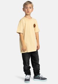 Beige T-shirt met korte mouwen met een kleurrijk hartlogo, gecombineerd met donkere jeans en grijze sneakers met aqua-accenten.