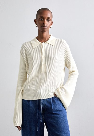 Cremefarvet ribbet sweater med krave og knaplukning, kombineret med løse blå denimbukser. Løs pasform, lange ærmer.