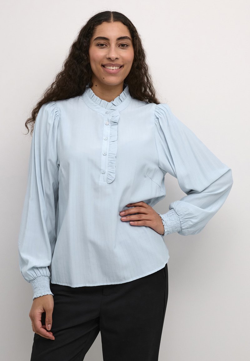 Blouse bleu clair avec de longues manches bouffantes, un col volanté et un plastron à boutons. Tissu lisse avec des rayures verticales et des poignets froncés.